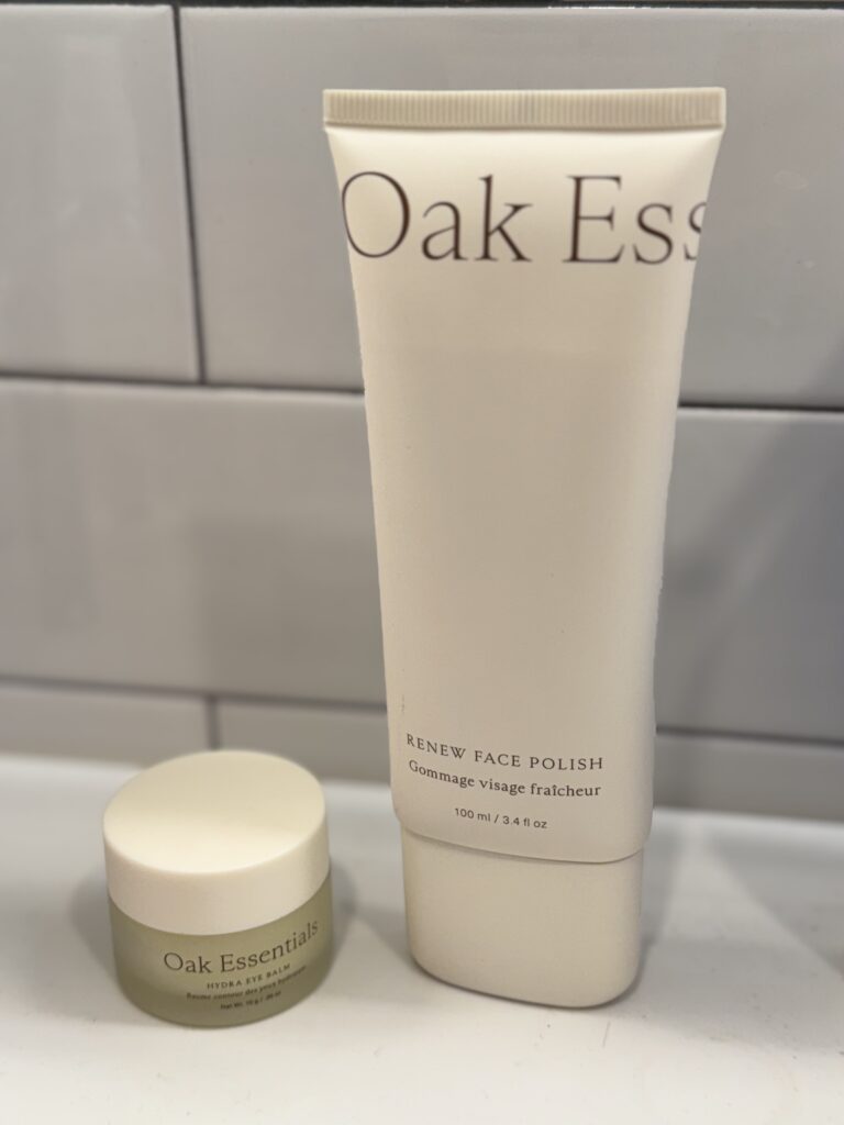 Two Oak Essentials skincare products—a tall white tube labeled 'Renew Face Polish' and a small beige jar labeled 'Oak Essentials'—on a white shelf.
