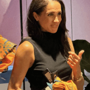 Woman with dark hair in a black sleeveless top, holding an orange garment and various items, looking to the side indoors.