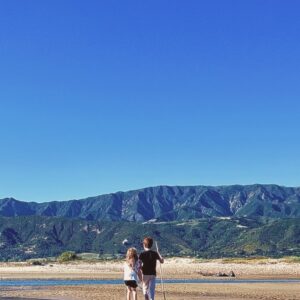 Two people walk along a sandy shore with a distant mountain range and clear blue sky behind them, one holding a stick.