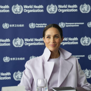 Woman in a pale lavender coat sits at a WHO backdrop with World Health Organization logos, smiling at the camera.