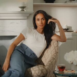 Woman relaxing on a patterned armchair in a cozy living room, running her dark hair with one hand and wearing a white tee and blue pants.
