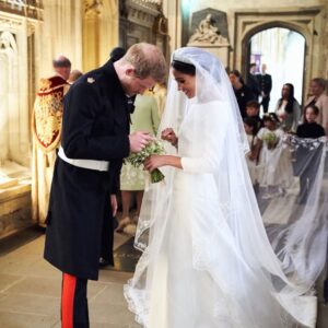 Bride in a white wedding gown and veil hands a small bouquet to a bowed military officer inside a stone church during a wedding.