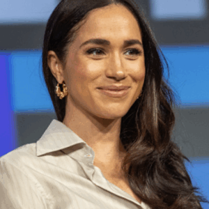 Portrait of a smiling woman with long dark hair, wearing a light blouse and gold hoop earrings, on a stage with a blue geometric backdrop.