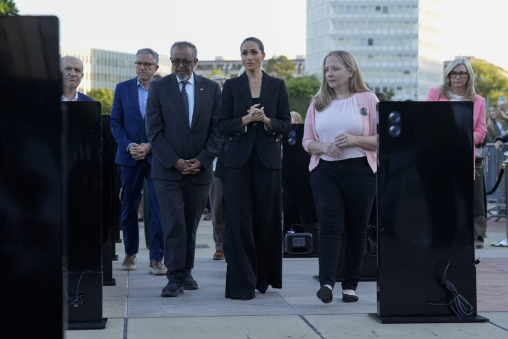 Group of formally dressed people walking outdoors between black display panels at an event. The woman in the center wears a black suit and leads the group.