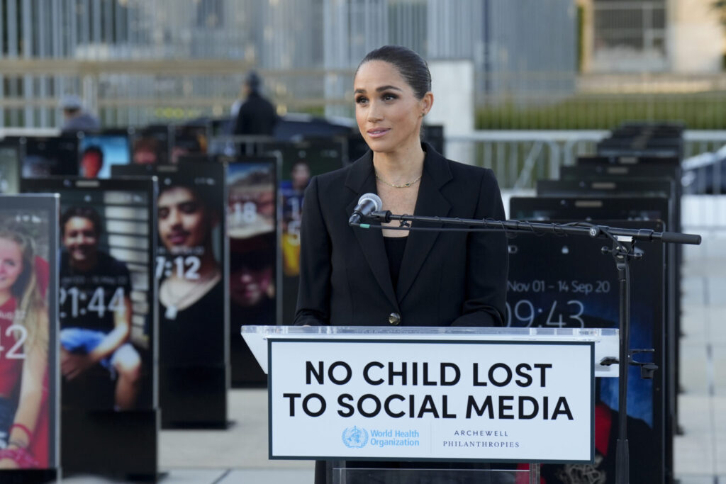Woman in a black blazer speaking at a podium with a sign that reads 'NO CHILD LOST TO SOCIAL MEDIA' outdoors.