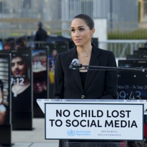 Woman in a black blazer speaking at a podium with a sign that reads 'NO CHILD LOST TO SOCIAL MEDIA' outdoors.