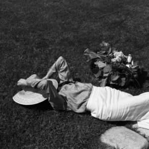 Person lying on a grassy lawn, wearing a striped shirt and wide-brim hat, with a bouquet and bottle placed nearby in a basket.