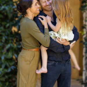 A smiling family of three hugging outdoors: mother in olive outfit, father in a dark shirt, and a young girl in a cream dress nestled between them.