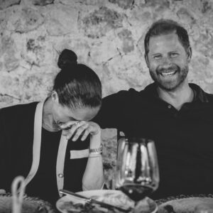 A smiling man with a beard sits beside a woman who covers her face in laughter at a dining table with a wine glass in the foreground, against a stone wall backdrop.