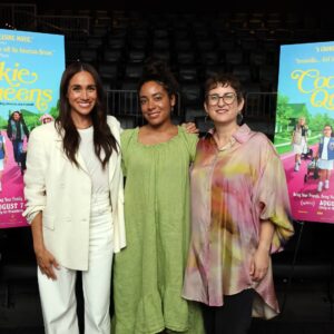 Three women smile for a photo at a movie premiere, standing in front of bright posters on either side; left wears a white suit, middle in a lime green dress, right in a pastel, tie-dye blouse.