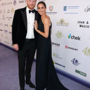 Couple posing on a purple carpet at a gala, man in a black tuxedo and bow tie, woman in a black evening gown beside him, backdrop covered with logos.