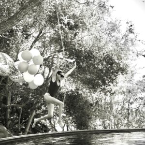 Woman in a black swimsuit leaps into a pool while holding a rope with white balloons tied to it, outdoors among trees (black-and-white photo).